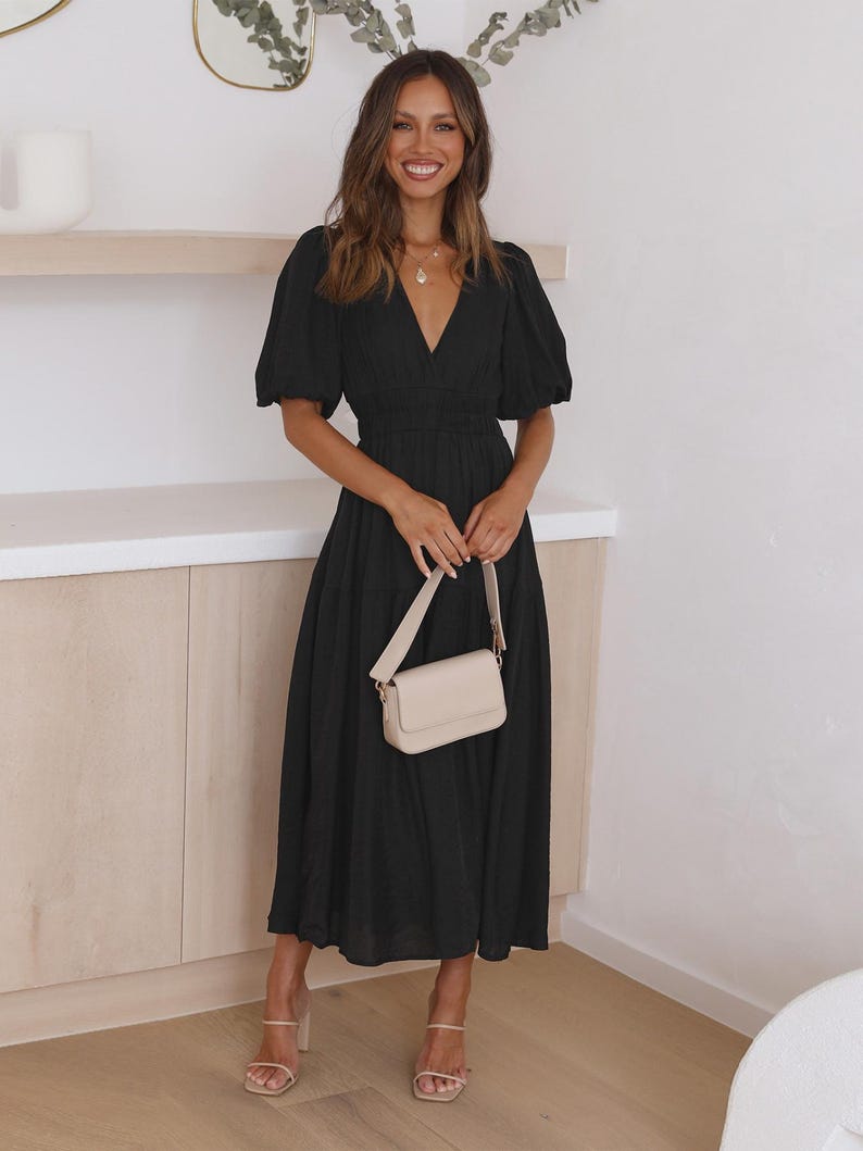 K&ouml;nnte beinhalten: Ein schwarzes Midikleid mit V-Ausschnitt und kurzen Puff&auml;rmeln. Das Kleid hat eine taillierte Taille und einen flie&szlig;enden Rock. Eine kleine, cremefarbene Handtasche mit Schulterriemen wird gehalten. Beigefarbene Abs&auml;tze vervollst&auml;ndigen den Look.