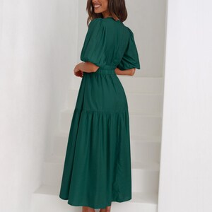 K&ouml;nnte beinhalten: Ein dunkelgr&uuml;nes Midikleid mit kurzen Puff&auml;rmeln und einer schmalen Taille. Das Kleid hat einen Stufenrock und wird mit nudefarbenen Sandalen mit Absatz getragen. Die Frau steht auf wei&szlig;en Treppen.