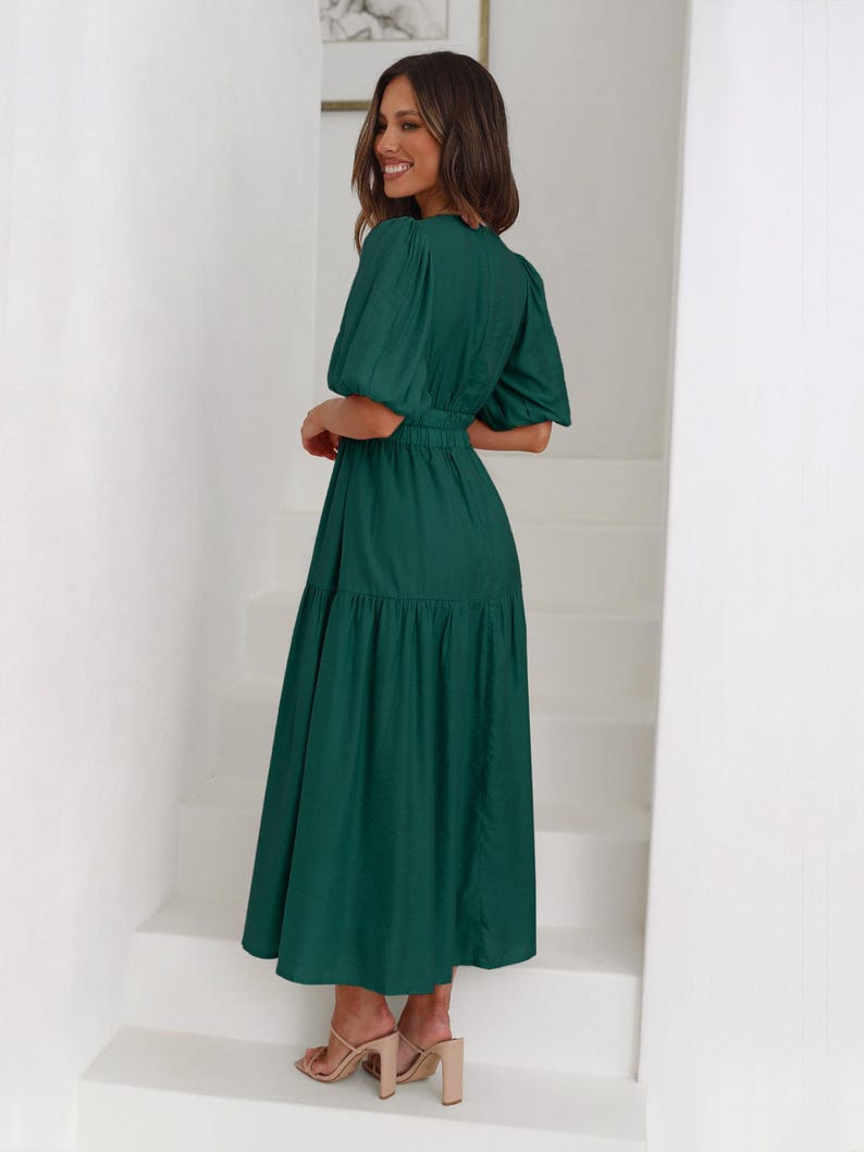 K&ouml;nnte beinhalten: Ein dunkelgr&uuml;nes Midikleid mit kurzen Puff&auml;rmeln und einer schmalen Taille. Das Kleid hat einen Stufenrock und wird mit nudefarbenen Sandalen mit Absatz getragen. Die Frau steht auf wei&szlig;en Treppen.