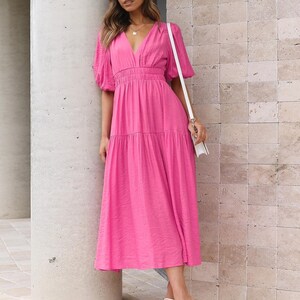 K&ouml;nnte beinhalten: Ein leuchtend pinkfarbenes Midikleid mit V-Ausschnitt und kurzen, gepufften &Auml;rmeln. Das Kleid hat einen Stufenrock und eine taillierte Taille. Die Person tr&auml;gt wei&szlig;e Sandalen und eine wei&szlig;e Tasche. Das Kleid ist aus strukturiertem Stoff.