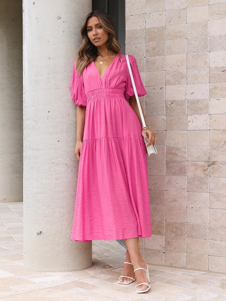K&ouml;nnte beinhalten: Ein leuchtend pinkfarbenes Midikleid mit V-Ausschnitt und kurzen, gepufften &Auml;rmeln. Das Kleid hat einen Stufenrock und eine taillierte Taille. Die Person tr&auml;gt wei&szlig;e Sandalen und eine wei&szlig;e Tasche. Das Kleid ist aus strukturiertem Stoff.