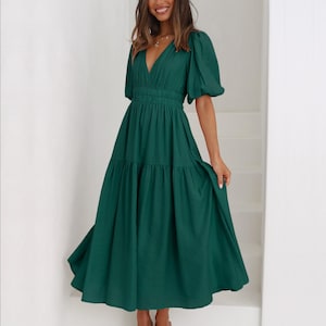 K&ouml;nnte beinhalten: Ein dunkelgr&uuml;nes Midikleid mit V-Ausschnitt und kurzen, gepufften &Auml;rmeln. Das Kleid hat eine taillierte Taille und einen Stufenrock. Das Model tr&auml;gt nudefarbene Sandalen. Das Kleid ist aus flie&szlig;endem Stoff.