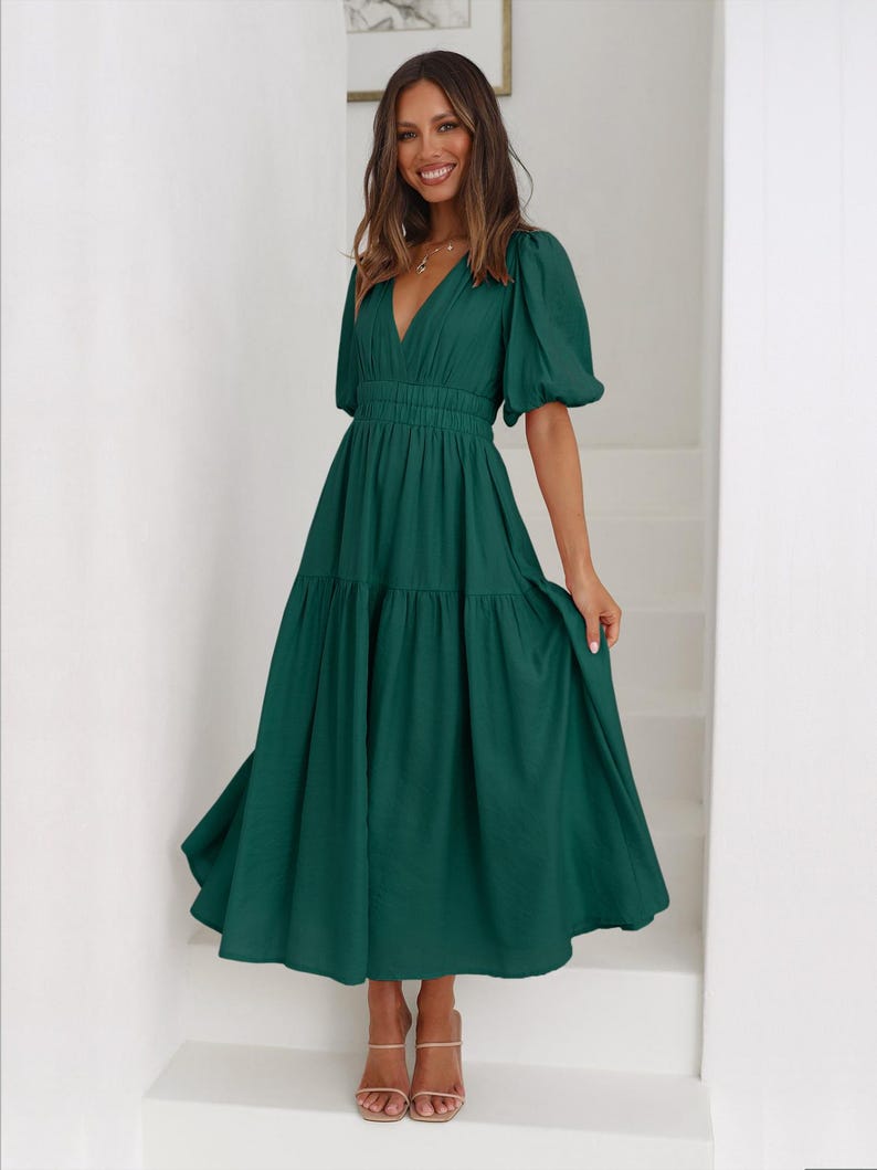 K&ouml;nnte beinhalten: Ein dunkelgr&uuml;nes Midikleid mit V-Ausschnitt und kurzen, gepufften &Auml;rmeln. Das Kleid hat eine taillierte Taille und einen Stufenrock. Das Model tr&auml;gt nudefarbene Sandalen. Das Kleid ist aus flie&szlig;endem Stoff.