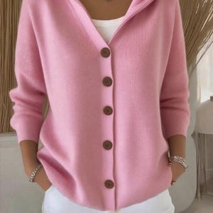 Könnte beinhalten: Ein hellrosa Kapuzen-Cardigan mit Knopfleiste. Der Cardigan hat lange Ärmel und wird mit einer weißen Hose getragen. Die Knöpfe sind braun, und der Stil ist lässig und bequem.