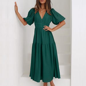 K&ouml;nnte beinhalten: Ein dunkelgr&uuml;nes Midikleid mit V-Ausschnitt und kurzen, gepufften &Auml;rmeln. Das Kleid hat eine taillierte Taille und einen Stufenrock. Das Model tr&auml;gt nudefarbene Sandalen. Das Kleid ist aus weichem, flie&szlig;endem Stoff.