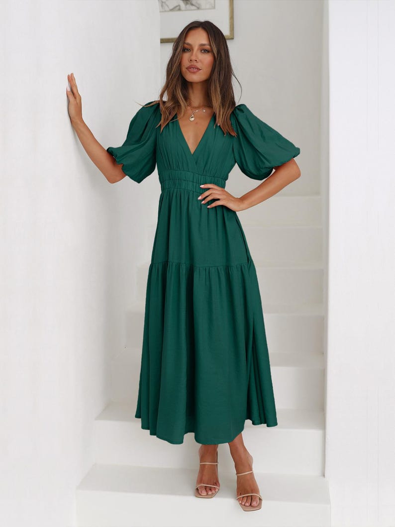 K&ouml;nnte beinhalten: Ein dunkelgr&uuml;nes Midikleid mit V-Ausschnitt und kurzen, gepufften &Auml;rmeln. Das Kleid hat eine taillierte Taille und einen Stufenrock. Das Model tr&auml;gt nudefarbene Sandalen. Das Kleid ist aus weichem, flie&szlig;endem Stoff.