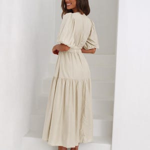 K&ouml;nnte beinhalten: Ein beiges Midikleid mit kurzen, gepufften &Auml;rmeln und einer schmalen Taille. Das Kleid hat einen Stufenrock und wird mit nudefarbenen Sandalen mit Absatz kombiniert. Die Frau steht auf einer wei&szlig;en Treppe.