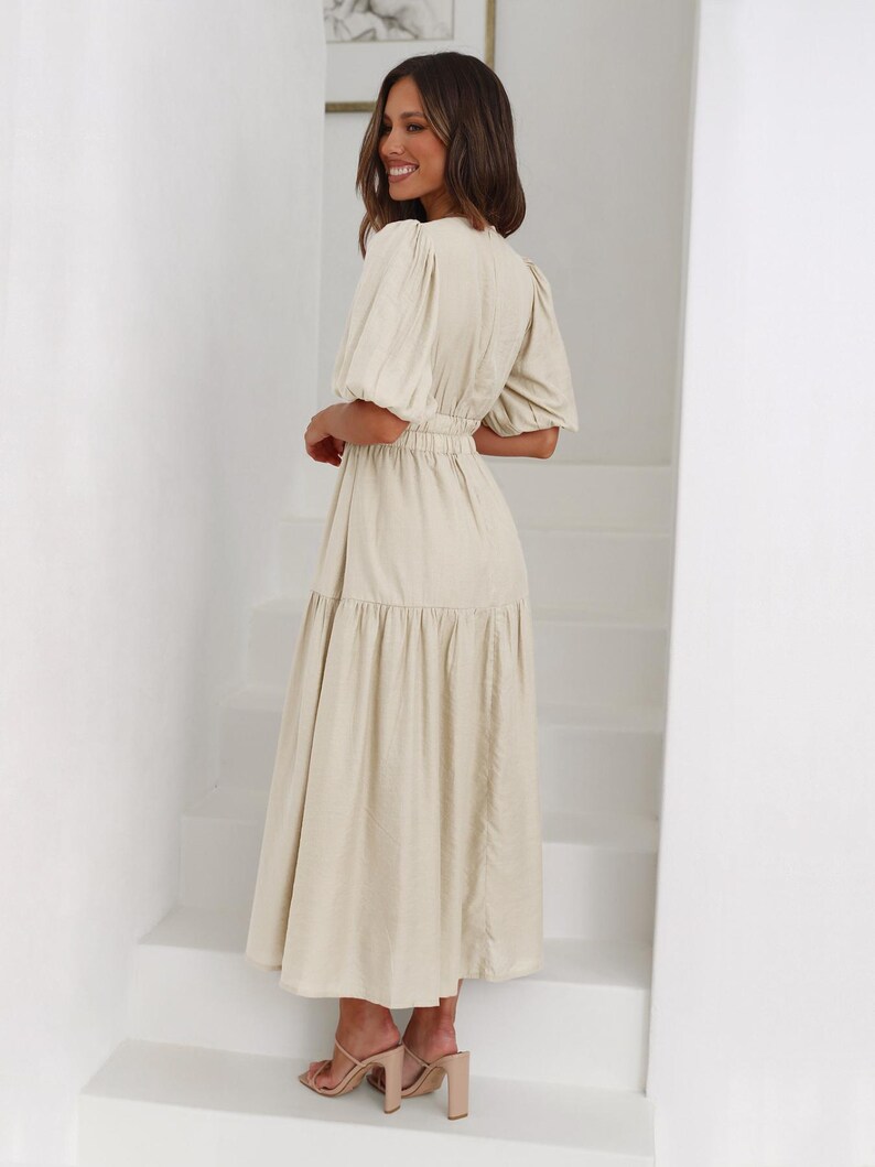 K&ouml;nnte beinhalten: Ein beiges Midikleid mit kurzen, gepufften &Auml;rmeln und einer schmalen Taille. Das Kleid hat einen Stufenrock und wird mit nudefarbenen Sandalen mit Absatz kombiniert. Die Frau steht auf einer wei&szlig;en Treppe.