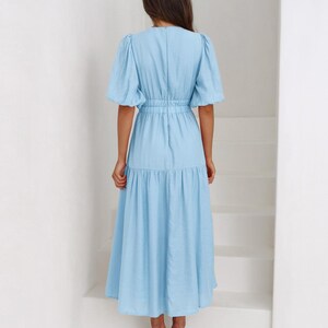 K&ouml;nnte beinhalten: Hellblaues Midikleid mit kurzen, gepufften &Auml;rmeln und einer schmalen Taille. Das Kleid hat einen Stufenrock und einen V-Ausschnitt. Das Model tr&auml;gt beige Abs&auml;tze. Das Kleid ist aus leichtem Stoff.