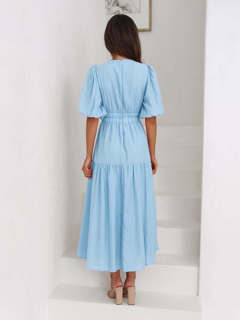 K&ouml;nnte beinhalten: Hellblaues Midikleid mit kurzen, gepufften &Auml;rmeln und einer schmalen Taille. Das Kleid hat einen Stufenrock und einen V-Ausschnitt. Das Model tr&auml;gt beige Abs&auml;tze. Das Kleid ist aus leichtem Stoff.