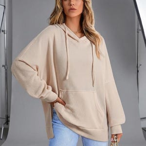 Könnte beinhalten: Beiger Kapuzenpullover mit Kängurutasche und Kordelzug. Das langärmlige Oberteil hat eine lockere Passform und wird mit hellblauen Jeans kombiniert. Das Outfit ist lässig und bequem.