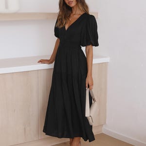 K&ouml;nnte beinhalten: Ein schwarzes Midikleid mit V-Ausschnitt und kurzen Puff&auml;rmeln. Das Kleid hat eine taillierte Taille und einen flie&szlig;enden Rock. Eine cremefarbene Handtasche und beige Abs&auml;tze vervollst&auml;ndigen das Outfit. Das Kleid ist f&uuml;r verschiedene Anl&auml;sse geeignet.