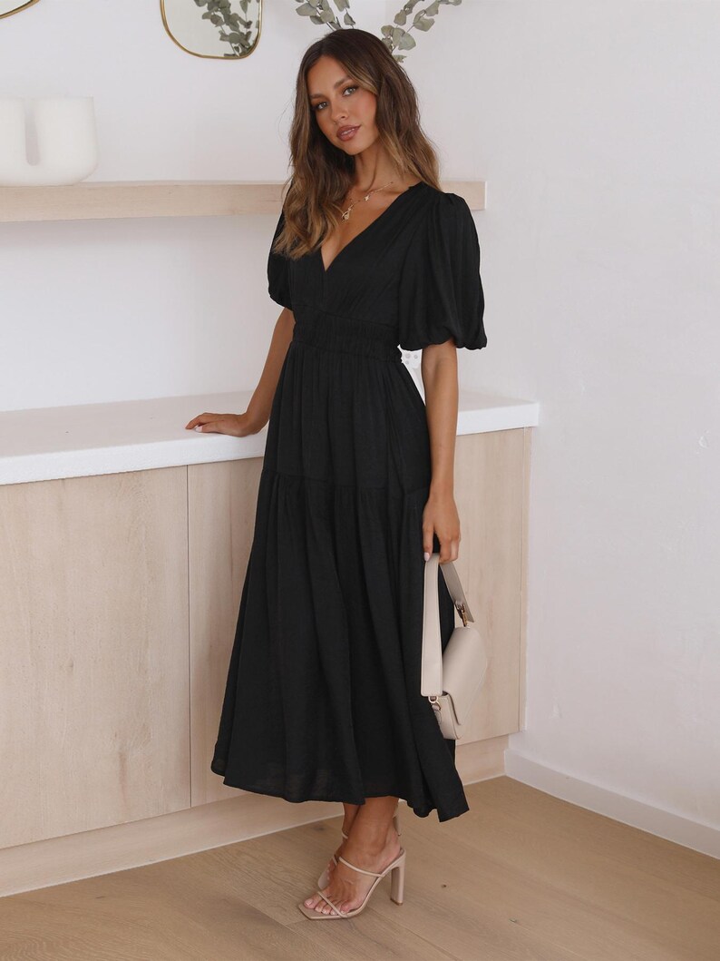 K&ouml;nnte beinhalten: Ein schwarzes Midikleid mit V-Ausschnitt und kurzen Puff&auml;rmeln. Das Kleid hat eine taillierte Taille und einen flie&szlig;enden Rock. Eine cremefarbene Handtasche und beige Abs&auml;tze vervollst&auml;ndigen das Outfit. Das Kleid ist f&uuml;r verschiedene Anl&auml;sse geeignet.