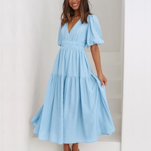 K&ouml;nnte beinhalten: Hellblaues Kleid mit V-Ausschnitt und kurzen Puff&auml;rmeln. Das Kleid hat eine taillierte Taille und einen Stufenrock. Das Model tr&auml;gt nudefarbene Abs&auml;tze. Das Kleid ist aus leichtem Stoff gefertigt.