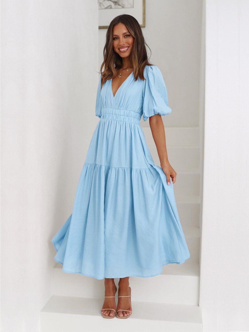 K&ouml;nnte beinhalten: Hellblaues Kleid mit V-Ausschnitt und kurzen Puff&auml;rmeln. Das Kleid hat eine taillierte Taille und einen Stufenrock. Das Model tr&auml;gt nudefarbene Abs&auml;tze. Das Kleid ist aus leichtem Stoff gefertigt.