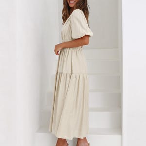 K&ouml;nnte beinhalten: Ein beiges Midikleid mit V-Ausschnitt und kurzen, gepufften &Auml;rmeln. Das Kleid hat eine taillierte Taille und einen Stufenrock. Das Model tr&auml;gt nudefarbene Sandalen mit Absatz. Der Hintergrund ist eine wei&szlig;e Treppe.