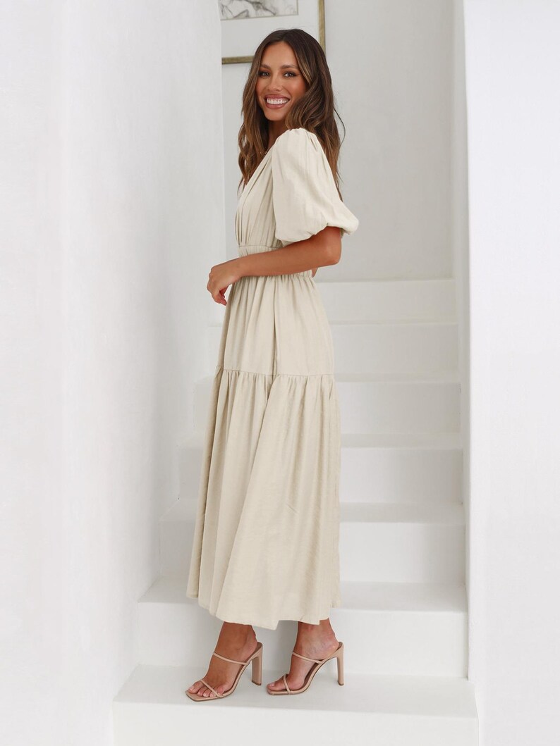 K&ouml;nnte beinhalten: Ein beiges Midikleid mit V-Ausschnitt und kurzen, gepufften &Auml;rmeln. Das Kleid hat eine taillierte Taille und einen Stufenrock. Das Model tr&auml;gt nudefarbene Sandalen mit Absatz. Der Hintergrund ist eine wei&szlig;e Treppe.