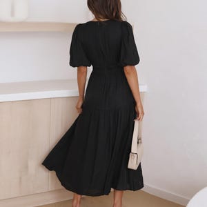 K&ouml;nnte beinhalten: Schwarzes Midikleid mit kurzen Puff&auml;rmeln und einer schmalen Taille. Das Kleid hat einen flie&szlig;enden Rock und wird mit beigen Abs&auml;tzen und einer passenden Handtasche getragen. Der Stoff des Kleides ist strukturiert.