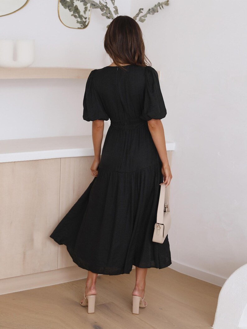 K&ouml;nnte beinhalten: Schwarzes Midikleid mit kurzen Puff&auml;rmeln und einer schmalen Taille. Das Kleid hat einen flie&szlig;enden Rock und wird mit beigen Abs&auml;tzen und einer passenden Handtasche getragen. Der Stoff des Kleides ist strukturiert.