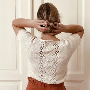 May include: Cream-colored knit top with short sleeves and a decorative openwork design down the back. The intricate pattern creates a textured look. The top is paired with rust-colored pants.