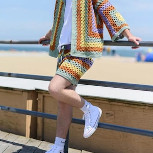 May include: A colorful crochet jacket and shorts set in shades of green, orange, yellow, and purple. The jacket has a collared neckline and three-quarter sleeves. The shorts are high-waisted. The outfit is paired with a white t-shirt and high-top sneakers.