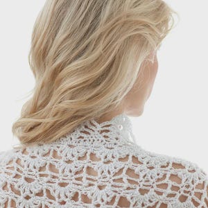 Può includere: Primo piano di un top bianco all'uncinetto con collo alto e motivo floreale. Il design a trama aperta è ben visibile. I capelli biondi del modello sono visti da dietro, con focus sul dettaglio all'uncinetto.