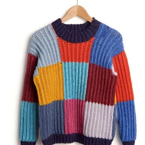 Op de afbeelding: Een kleurrijke patchwork trui met een geribbelde textuur, tentoongesteld op een houten hanger. De trui heeft een verscheidenheid aan kleuren, waaronder blauw, oranje, rood, geel en roze, gerangschikt in een geometrisch patroon. De halslijn en manchetten zijn donker marineblauw.