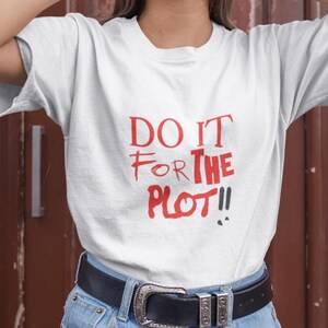 Puede incluir: Camiseta blanca con la frase roja "DO IT FOR THE PLOT!!". La camiseta se combina con vaqueros azules y un cinturón negro con hebilla plateada. El texto está escrito a mano.
