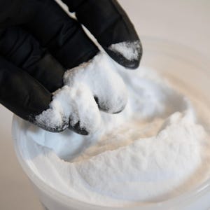 May include: Close-up of a hand in a black glove holding a clump of white powder above a container filled with the same powder. The powder appears fine and granular, with a bright white color. The background is a soft, neutral tone.