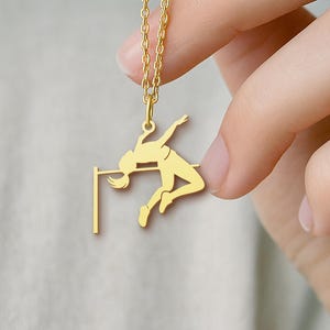May include: A gold-colored pendant necklace featuring a high jumper clearing a bar. The pendant is a silhouette of an athlete in mid-air. The necklace is held by a person.