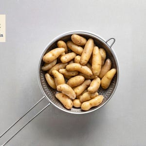 May include: A stainless steel colander filled with small, oblong, light brown potatoes. The colander has a long handle and is set against a light gray background. The OliveJardin logo is in the upper left corner.