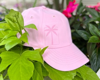 May include: A light pink baseball cap with a palm tree graphic. The cap has a curved brim and small ventilation holes. The hat is surrounded by green foliage and flowers.
