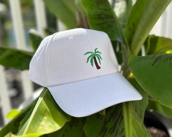 May include: A white baseball cap with a curved brim. The cap features an embroidered palm tree design in green and brown on the front. The hat is set against a backdrop of green leaves.