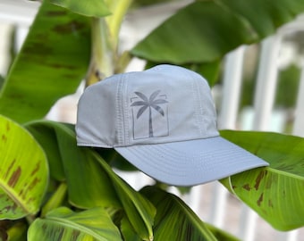 May include: A light gray baseball cap featuring a palm tree graphic within a rectangular frame on the front. The cap is set against a backdrop of large green and brown banana leaves, creating a tropical vibe.