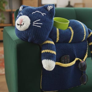 Puede incluir: Organizador de sofá con forma de gato tejido en azul marino con rayas y detalles amarillos. El gato tiene cara y patas blancas y sostiene una taza verde. Bolsillos para mando a distancia y gafas.