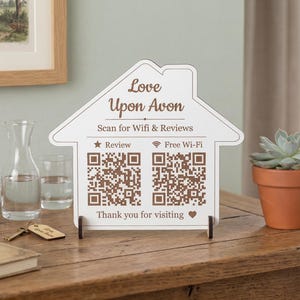 May include: A white sign in the shape of a house, with the words "Love Upon Avon" and QR codes for Wi-Fi and reviews. It stands on a wooden table, next to a small potted succulent and books.