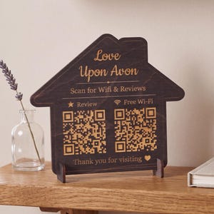 Op de afbeelding: Een donkerbruin houten bord in de vorm van een huis met gouden tekst en twee QR-codes. De tekst luidt Love Upon Avon, Scan for Wifi & Reviews, Review, Free Wi-Fi en Thank you for visiting. Het bord staat op twee kleine houten voetjes.