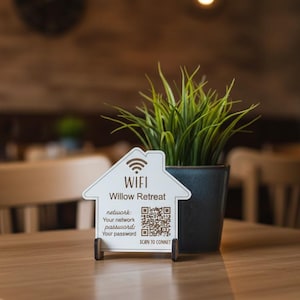 Peut inclure: Un panneau blanc en forme de maison avec le mot "WIFI" et le texte "Willow Retreat" imprimés dessus. Le panneau affiche également les informations réseau et un code QR. Une petite plante en pot avec de l'herbe verte est derrière le panneau sur une table en bois.