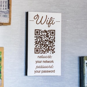 May include: A white rectangular magnet with the word "Wifi" in a decorative font, a QR code, and network information. The magnet is attached to a silver fridge door, alongside other decorative magnets.