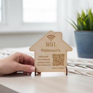 May include: Wooden house-shaped WiFi sign with the text "WIFI Robinson's" and a QR code. The sign also displays network information and a "SCAN TO CONNECT" prompt. The sign is on a table with a plant in a blue pot in the background.