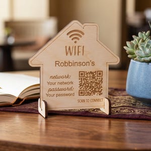 May include: A light-colored wooden sign shaped like a house, featuring an engraved WiFi symbol, the text "Robinson's", "network: Your network", "password: Your password", and "SCAN TO CONNECT" alongside a QR code, supported by a small wooden stand.