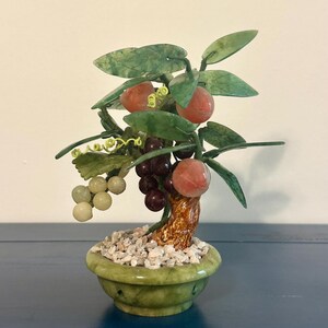 May include: A decorative jade bonsai tree with green leaves, red and purple fruit, and a brown trunk. The tree sits in a green pot filled with small white stones. A unique home decor item.