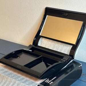 May include: Black compact makeup case with a built-in mirror and two small compartments. The case is open, revealing the mirror and the empty compartments. The case is sitting on top of a black box with text.