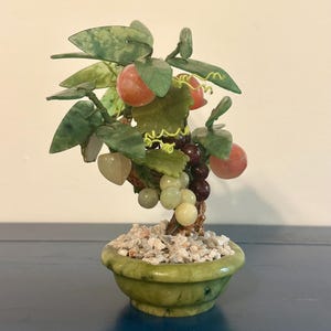 May include: A small, decorative jade bonsai tree with green leaves, red-orange fruit, and clusters of grapes in a green pot filled with small white stones. The tree is a miniature sculpture.