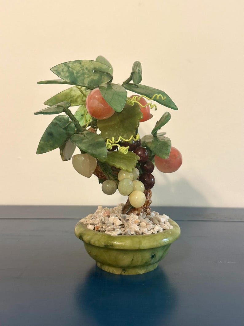 May include: A small, decorative jade bonsai tree with green leaves, red-orange fruit, and clusters of grapes in a green pot filled with small white stones. The tree is a miniature sculpture.
