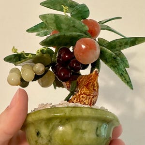 May include: A small, decorative jade bonsai tree with green leaves, red and purple fruit, and a light green pot. The tree is held in a hand, showcasing the intricate details of the handcrafted piece.