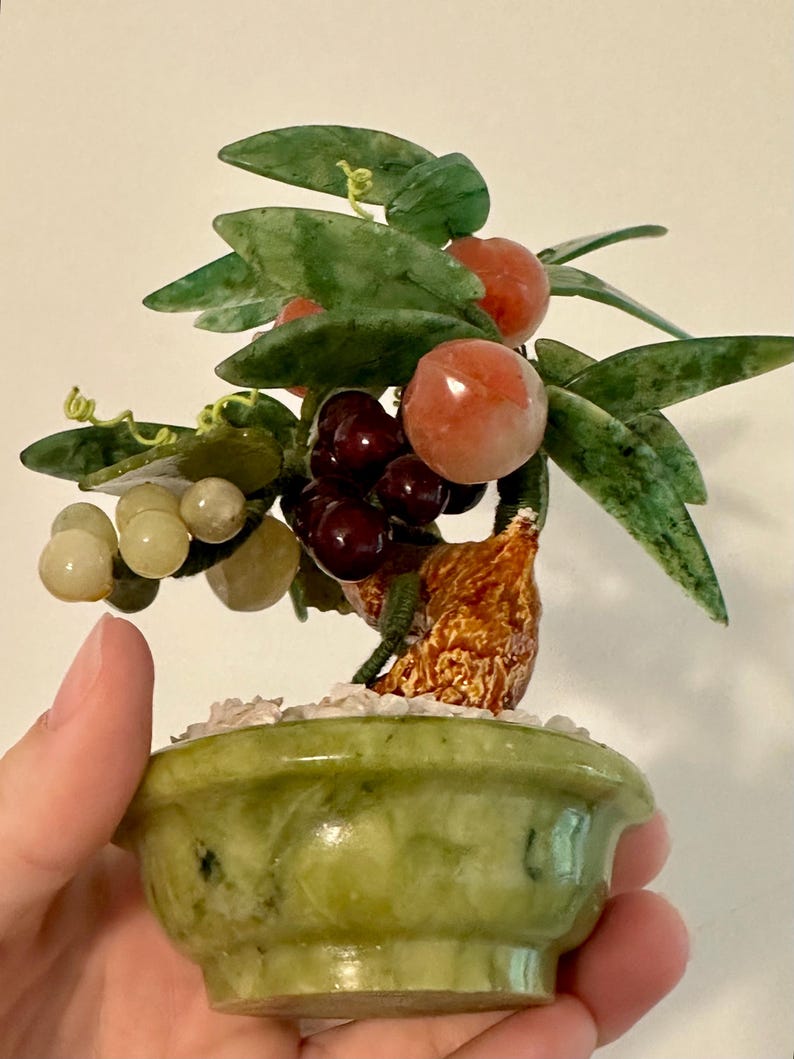 May include: A small, decorative jade bonsai tree with green leaves, red and purple fruit, and a light green pot. The tree is held in a hand, showcasing the intricate details of the handcrafted piece.