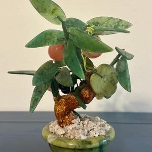 May include: A jade bonsai tree sculpture with green leaves and small orange fruit. The tree is in a green pot filled with small white stones. The trunk is a textured brown color.