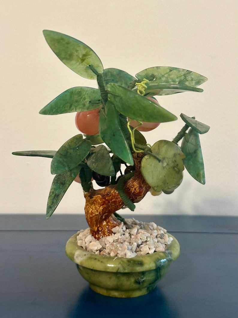 May include: A jade bonsai tree sculpture with green leaves and small orange fruit. The tree is in a green pot filled with small white stones. The trunk is a textured brown color.