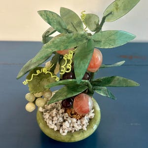 May include: A small, decorative jade tree with green leaves, red-orange fruit, and clusters of grapes. The tree is potted in a round, green container filled with small white stones. The background is a blue surface.